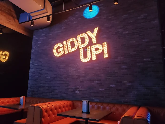 Lit-bulb GIDDY UP! marquee sign on a brick wall above orange tufted booths